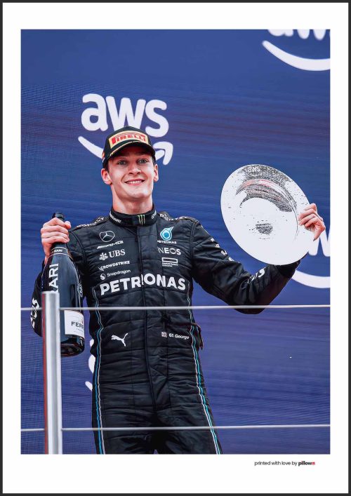 George Russell na pódiu Formuly 1, drží šampanské a víťazný pohár, v pretekárskom obleku, s reklamnými bannermi v pozadí.